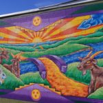 A colorful mural on a brick wall shows a landscape with a river, a stone bridge, green hills, a sunrise, and animals including goats, a cow, and a rabbit. The artwork features bold outlines and vibrant colors.