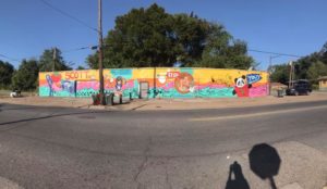A long mural on a yellow wall along a street features colorful designs including a panda, fish, a stop sign, and the word SCOTT in red letters, with trees and a sidewalk in the background.