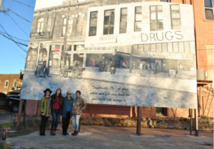 Four people stand in front of a large mural depicting a historic street scene with old cars and storefronts, including signs for “DRUGS” and “T.S.D. McKINNEY,” painted in sepia tones.