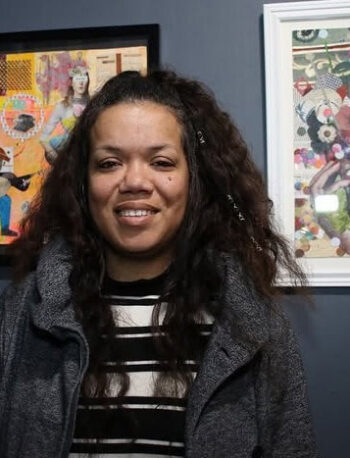A woman with long curly hair stands in front of a dark wall with two colorful framed artworks. She wears a striped shirt and a gray jacket, and is smiling slightly at the camera.
