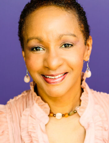 A woman with short curly hair, wearing drop earrings and a beaded necklace, smiles at the camera. She is dressed in a peach ruffled top and is posed in front of a plain purple background.