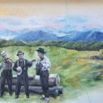 A mural depicts three musicians in old-fashioned clothes playing a fiddle, banjo, and guitar in front of logs, with rolling hills, a cabin, trees, and mountains in the background under a pastel sky.