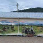 A mural on a long building wall depicts people inside with vintage equipment, transitioning to an outdoor scene with cattle, trees, and mountains under a cloudy sky, blending indoor and rural landscapes.