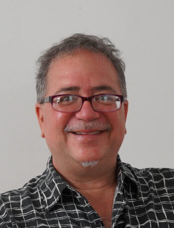 A man with short gray hair and glasses smiles at the camera. He is wearing a black shirt with a white grid pattern. The background is plain and light-colored.