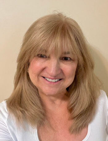 A woman with shoulder-length blonde hair and bangs smiles at the camera. She is wearing a white top and is standing against a plain beige background.