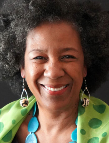 A woman with curly gray hair smiles at the camera. She wears patterned earrings, a green scarf with polka dots, and a blue necklace, with a plain dark background behind her.