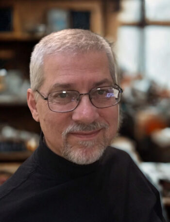 A middle-aged man with short gray hair, a goatee, and glasses is wearing a black turtleneck and smiling slightly. The background is softly blurred and suggests an indoor, possibly home or office, setting.