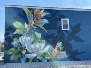 A mural painted on a blue wall depicts white magnolia flowers and green leaves. There is a single window on the upper right side, and sunlight creates a lens flare across the image. White gravel covers the ground below.