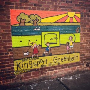 A mural on a brick wall depicts children and a dog walking and playing in front of a park scene with trees, swans, a squirrel, and a sunrise. The words Kingsport Greenbelt are written at the bottom.