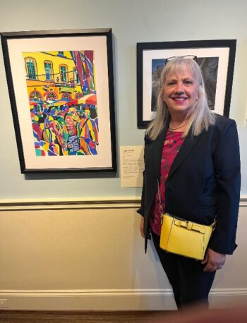 A woman with gray hair, wearing a dark blazer, pink top, and yellow purse, stands indoors next to a colorful framed painting and a black-and-white photo displayed on a light-colored wall.