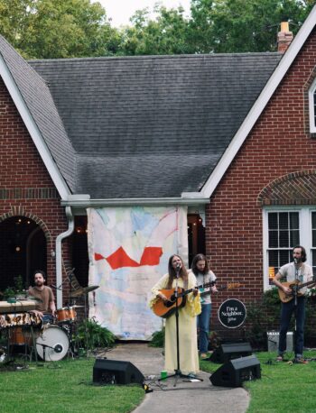 A band performs outdoors in front of a brick house. Three musicians play guitar and sing, while two play keyboard and drums. Sound equipment and a large abstract backdrop are set up on the front lawn.