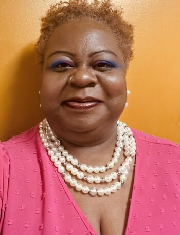 A woman with short curly hair, wearing vibrant blue eye makeup, a pink top, and layered pearl necklaces, stands in front of an orange wall, smiling at the camera.