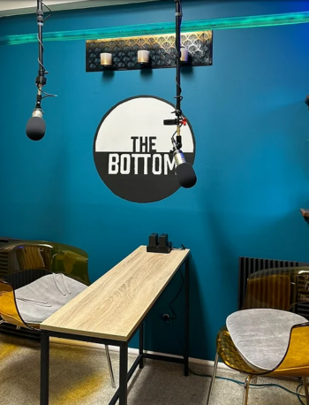 A podcast studio with a wooden table, two transparent chairs with gray seat cushions, two hanging microphones, a teal wall with The Bottom logo, shelves with plants, and a black radiator.