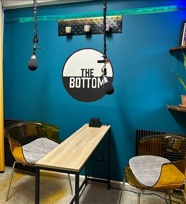 A podcast studio with a wooden table, two transparent chairs with gray seat cushions, two hanging microphones, a teal wall with The Bottom logo, shelves with plants, and a black radiator.