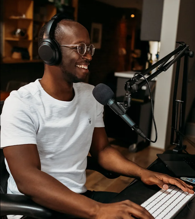 A person wearing headphones and glasses sits at a desk, smiling while speaking into a microphone and typing on a keyboard. A computer monitor and microphone are visible in a home office setting.