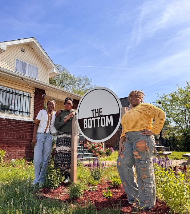 Three people stand outdoors next to a circular sign that reads “THE BOTTOM” in front of a brick house with a white roof. The yard has green grass and flowers. The sky is clear and blue.