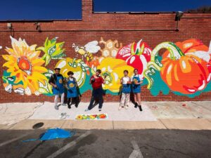 Five people stand in front of a brick wall painted with a colorful mural featuring large flowers, bees, and a pumpkin. They are holding painting supplies and wearing blue aprons. The ground is covered with tarps.