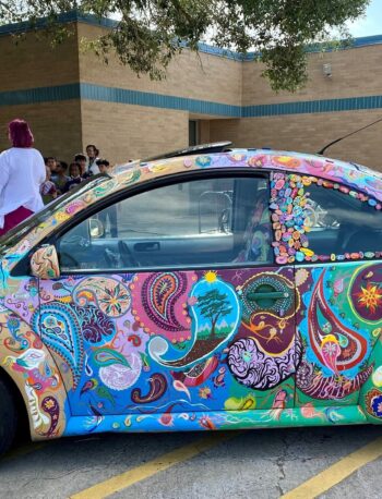 A Volkswagen Beetle decorated with colorful paisley and floral patterns is parked outside a brick building. The car’s wheels have orange spokes. People stand and sit in the background near the building.