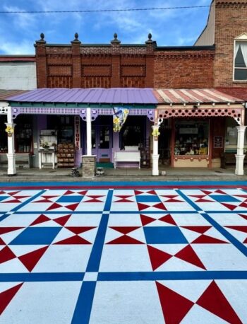 A street scene shows a colorful geometric mural painted on the road in front of historic brick and wood storefronts, featuring red, blue, and white patterns under a partly cloudy sky.