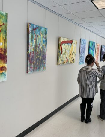 Two people walk down a hallway with abstract paintings displayed on the wall; the hallway is well-lit with a white tile floor and ceiling lights.