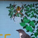 A mural painted on a wall shows a large spider on a web above green leaves, with a bird below on a blue background.