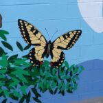 A mural of a yellow and black butterfly with red spots on its lower wings, perched on green leaves, painted on a blue cinder block wall with part of a white shape in the background.