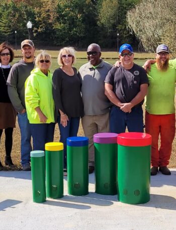 A group of twelve adults stands outdoors in a row behind colorful recycling bins near a lake, with trees in the background and two informational signs on either side of the bins.