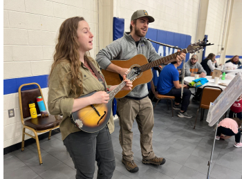 A woman playing a mandolin and a man playing a guitar perform together indoors. Several people are seated at tables in the background, and an empty chair with water bottles sits to the side.