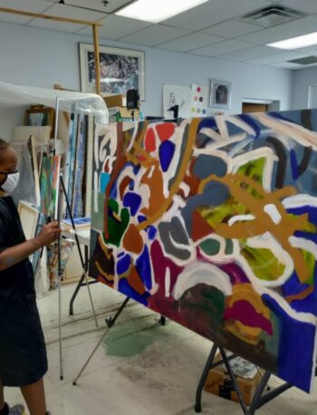 A person wearing an apron and mask stands in an art studio, painting a large abstract canvas with various bright colors and shapes. The studio is filled with art supplies, canvases, and paintings.