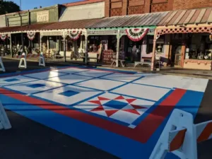 A painted quilt pattern featuring geometric shapes and a red star is on a street in front of old-fashioned storefronts decorated with patriotic bunting and vintage items. Traffic barricades block off the area.