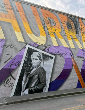 A mural on a brick wall reads HURRAH VOTE in large letters, with black script writing over it. There is also a black-and-white photograph of a woman wearing glasses and a suit.