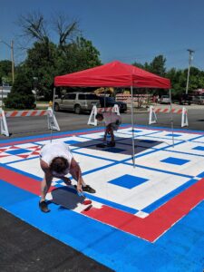 Two people paint a large geometric pattern on pavement under a red canopy on a sunny day. Traffic barricades and parked vehicles are visible in the background. One person rolls red paint on the ground in the foreground.