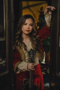 A woman with long wavy hair stands in a doorway, wearing a red and gold embroidered jacket over a beige blouse. She is holding a mandolin vertically and looks directly at the camera with a neutral expression.