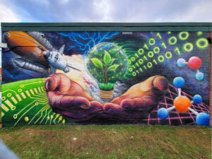 A mural shows hands holding a light bulb with a growing plant, surrounded by a rocket, circuit board, binary code, molecules, and lightning on a brick wall under a cloudy sky.