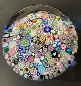 A glass paperweight with intricate, colorful millefiori patterns arranged in concentric circles, featuring various shapes and designs on a black surface background.