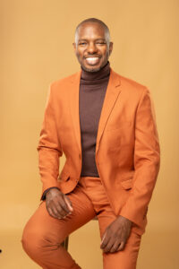 A man wearing an orange suit and brown turtleneck sits on a stool against a plain beige background, smiling at the camera.