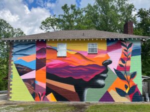 A vibrant mural on the side of a house depicts a stylized profile of a persons face filled with colorful mountain and sunset scenes, geometric shapes, and plant motifs against a yellow and green background.