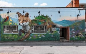 A mural on a brick building features a fox, a bird, and a deer surrounded by plants, flowers, and mountains. The mural is signed “Ernesto Maranje.” There are two sets of windows and an open doorway in the wall.