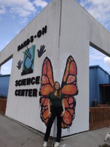 A person stands in front of a white building labeled Hands-On Science Center, positioned at the corner where orange butterfly wings are painted on the wall, appearing as if they have wings.