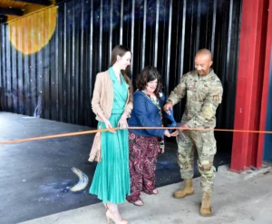 Three people, including a man in military uniform, use large scissors to cut an orange ribbon indoors. The backdrop features a space-themed mural with planets and stars.