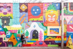 A person leans against a colorful mural wall featuring abstract shapes, a bear, a lion, plants, mountains, and a blue ladder. The mural includes bright colors and geometric patterns. Overhead wires are visible.