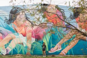 A person in a gray hoodie stands in front of a large mural featuring three girls in colorful outfits, painted on a wall, with tree branches partially obscuring the view.