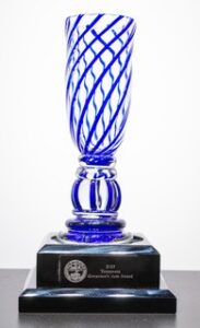 A blue and clear glass trophy with diagonal stripes sits on a black base. The base has a silver plaque that reads 2023 Governor’s Arts Award along with an official seal.