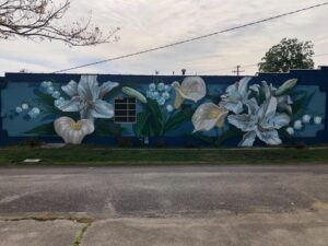 A mural painted on a blue wall depicts large white lilies, calla lilies, and lily of the valley flowers with green leaves. There is a single window in the mural, and the scene is outdoors along a quiet street.