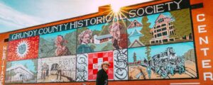 A mural on a red brick building reads Grundy County Historical Society and Heritage Center, featuring painted scenes of local history, people, buildings, and a train. A person walks by in front of the mural.