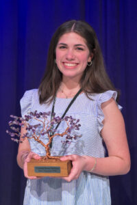 A young woman with long brown hair, wearing a light blue and white striped dress, smiles while holding a trophy shaped like a tree, standing in front of a dark blue curtain.
