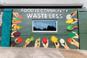 A mural on a brick wall featuring the words FOOD IS COMMUNITY WASTE LESS with hands holding various fruits and vegetables painted around a central window.