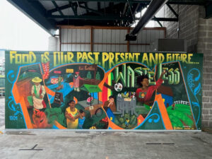 A colorful mural shows various people, food items, farms, cityscapes, and the words Food is our past, present and future... Waste less. The artwork blends rural and urban scenes under a covered outdoor area.