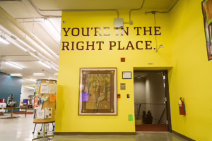 A yellow wall with large text reading YOURE IN THE RIGHT PLACE. Below is a framed artwork and several plaques. To the left is a bulletin board, and to the right is an open doorway leading to a stairwell.