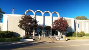 A two-story public library building with three large, arched entryways, wide steps leading up to the entrance, two trees flanking the front doors, and an American flag on a pole to the right.
